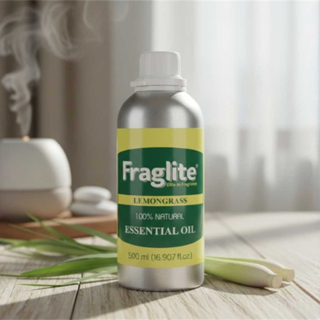 A 500 ml aluminum bottle of Fraglite Lemongrass Essential Oil on a wooden table with fresh lemongrass stalks and an active oil diffuser in the background.
