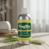 A 500 ml aluminum bottle of Fraglite Lemongrass Essential Oil on a wooden table with fresh lemongrass stalks and an active oil diffuser in the background.