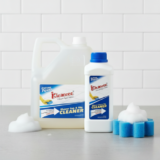 A large clear gallon jug and a smaller white bottle of Kleanzee Shower Tub & Tile Cleaner standing on a grey surface next to a blue sponge and white cleaning foam.
