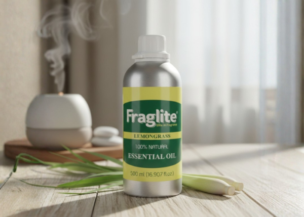 A 500 ml aluminum bottle of Fraglite Lemongrass Essential Oil on a wooden table with fresh lemongrass stalks and an active oil diffuser in the background.