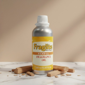 A silver Fraglite bottle with a yellow "Sandal Fragrance Oil" label, centered on a white marble surface surrounded by pieces of natural sandalwood.