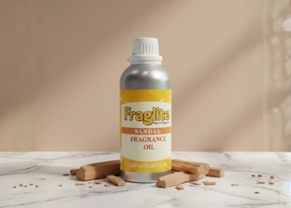 A silver Fraglite bottle with a yellow "Sandal Fragrance Oil" label, centered on a white marble surface surrounded by pieces of natural sandalwood.