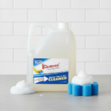 A large, clear plastic gallon jug with a white handle and Kleanzee label, featuring cleaning foam and a blue flower-shaped sponge on a marble countertop.