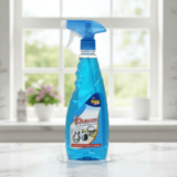 A clear spray bottle filled with blue Kleanzee glass cleaner liquid, standing on a marble countertop with a blurred window and garden background.