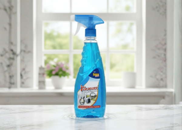 A clear spray bottle filled with blue Kleanzee glass cleaner liquid, standing on a marble countertop with a blurred window and garden background.