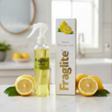 A Fraglite Lemon air freshener spray and box on a marble surface with sliced and whole lemons in a bright kitchen or bathroom background.