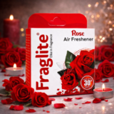 A box of Fraglite Rose Air Freshener placed among red roses and lit tea light candles in a warm, dimly lit environment.